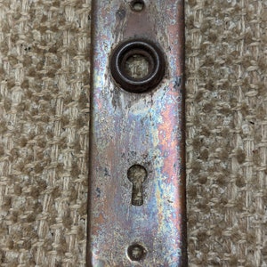 Peut inclure: Une plaque de porte rectangulaire vintage avec un trou de serrure et un bouton rond. La plaque est en métal et présente une patine usée et vieillie avec des nuances de cuivre, de bleu et de vert.