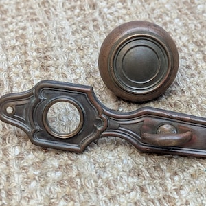 May include: Antique brass door knob and backplate with a decorative design. The knob has a raised center and a ring around it.