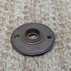 May include: A circular, dark brown metal mounting plate with a raised, stepped center ring and two small screw holes on opposite sides. The surface has a weathered, textured finish with visible dark oxidation around the central opening.