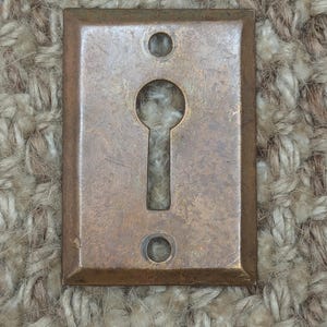 Op de afbeelding: Een messing sleutelgat afdekplaat met een rechthoekige vorm en twee ronde gaten aan weerszijden van het sleutelgat.