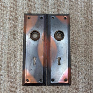 May include: Two rectangular metal door plates featuring a dark, weathered finish with copper-toned highlights. Each plate includes a central circular opening for a knob, a lower keyhole, and four small screw holes at the corners for mounting.