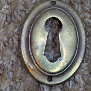 Pode incluir: Uma tampa de buraco de fechadura de latão com forma oval e uma borda decorativa. A tampa tem dois pequenos furos na parte superior e inferior.