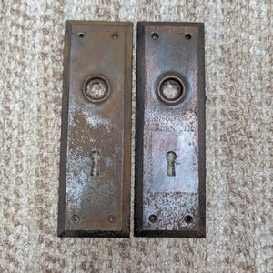 Könnte beinhalten: Zwei rechteckige Türschilder aus Metall mit einer verwitterten, rostbraunen Oberfläche. Jedes Schild verfügt über eine kreisförmige Mittelöffnung für einen Türgriffdorn und einen unteren Schlüssellochausschnitt. Vier kleine Schraublöcher sind vorhanden.