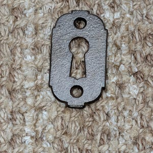 Könnte beinhalten: Dekorative, metallgraue Schlüssellochabdeckung mit strukturierter Oberfläche. Die Abdeckung hat eine klassische, rechteckige Form mit abgerundeten Kanten und zwei kleinen Löchern über und unter der Schlüssellochöffnung. Das Schlüsselloch hat eine traditionelle Tropfenform.