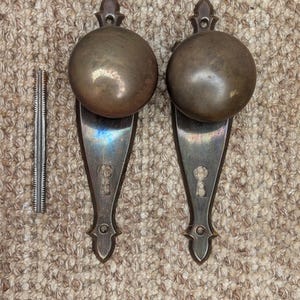 May include: Two antique brass door knobs with keyholes and ornate detailing. The knobs are round and have a slightly aged finish. A small metal rod is included.