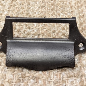 May include: A black metal label holder with a rectangular opening and two screw holes. The holder has a textured surface and a curved bottom edge. Ideal for drawers and cabinets.