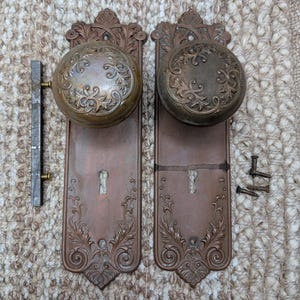 May include: Antique bronze door hardware set, featuring two ornate doorknobs with detailed floral patterns. The set includes matching backplates with keyholes and decorative scrollwork, plus a separate latch and screws.