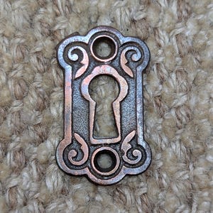 May include: A vintage keyhole cover made of metal with a decorative design. The cover is a dark brown color with a copper patina. It has a keyhole in the center and two decorative circles on either side.