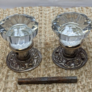 May include: Two antique glass door knobs with ornate metal bases and a single screw. The knobs are clear glass with a faceted design.