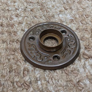 Può includere: Un rosone per porta circolare in metallo color bronzo con un motivo a volute in rilievo e un collare cilindrico centrale. L'accessorio presenta tre fori di montaggio equidistanti lungo il perimetro per l'installazione su una porta.