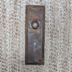 Op de afbeelding: Een rechthoekige metalen deurplaat met een verweerde bronskleurige afwerking. De plaat heeft een centrale ronde opening voor een deurklink, een sleutelgat eronder en vier kleine schroefgaten in de hoeken van het verticale frame.