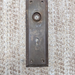 Op de afbeelding: Een rechthoekige, verweerde bruine metalen deurplaat met een afgeschuinde rand, een centrale ronde opening voor een deurknop en een onderste sleutelgat. De plaat heeft vier kleine schroefgaten in de hoeken voor montage.