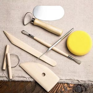 Könnte beinhalten: Eine Sammlung von Töpferwerkzeugen, darunter ein Metall-Nierenwerkzeug, ein Schleifwerkzeug, eine Holzrippe, ein Nadelwerkzeug und ein gelber Schwamm. Die Werkzeuge sind auf einem neutralfarbenen Stoff angeordnet, ideal zum Formen von Ton.