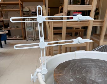 May include: White pottery wheel tool with adjustable arms and blades. The tool is attached to a pottery wheel with a clamp.