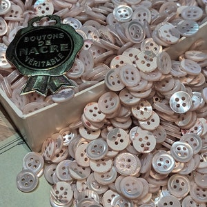 May include: A large pile of white mother of pearl buttons with a pink iridescent sheen. The buttons are in a white box with a gold seal that reads "Boutons de Nacre Veritable".