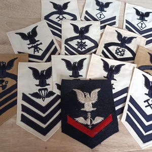 Könnte beinhalten: Eine Sammlung von Vintage-Militärabzeichen mit verschiedenen Designs. Die Abzeichen zeigen dunkelblaue und beige Farbtöne mit gestickten Adlern und Chevron-Mustern. Einige Abzeichen haben zusätzliche Symbole wie Anker und Propeller.