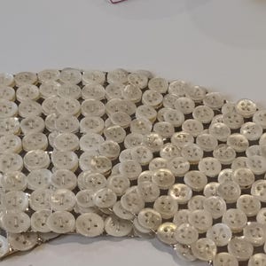May include: A collection of small, off-white buttons arranged closely together. The buttons are round with four holes and are attached to a silver-colored backing. The arrangement forms a curved shape.
