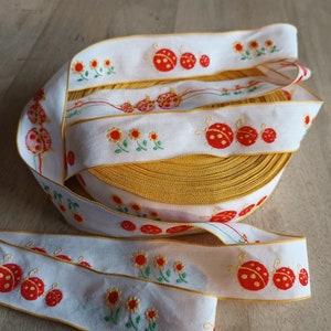 Może przedstawiać: Biała wstążka z żółtą obwódką i haftowanymi czerwonymi biedronkami i żółtymi kwiatami.