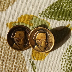 Peut inclure: Deux boutons en métal couleur or avec un profil de Napoléon Bonaparte portant un bicorne. Les boutons portent l'inscription "NAPOLEON" sur le bord.
