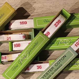 May include: A collection of vintage green and yellow boxes, each containing white measuring tape with red numbers. The boxes are labeled with French text, including "Une Grosse Garantie" and "Yester Coton Made in France."