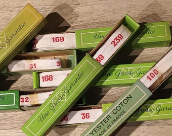 May include: A collection of vintage green and yellow boxes, each containing white measuring tape with red numbers. The boxes are labeled with French text, including "Une Grosse Garantie" and "Yester Coton Made in France."