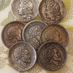 Peut inclure: Une collection de sept pièces de monnaie argentées représentant des personnages historiques différents. Les pièces ont des bords surélevés et des gravures détaillées des visages et des noms des personnages. Les pièces sont disposées en cercle sur une surface texturée.
