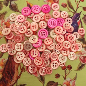 Pode incluir: Uma coleção de botões rosa em várias tonalidades, do rosa claro ao fúcsia vibrante, espalhados sobre um fundo com padrão floral. Os botões têm quatro furos e são feitos de material brilhante. O fundo apresenta um design floral verde com pássaros e flores.