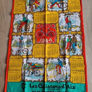 Puede incluir: Una colorida toalla de cocina vintage con un calendario para el año 1984. La toalla presenta ilustraciones de bicicletas y el texto "Les Calissons d'Aix" con el nombre de la empresa "Leonard Parli" y el número de teléfono "26.05.71".
