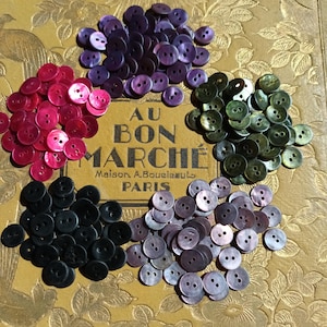Peut inclure: Une collection de boutons ronds à deux trous disposés en cinq groupes de couleurs distincts : violet foncé, rose vif, vert forêt, noir et lavande irisé. Les boutons sont présentés sur une surface dorée avec le texte en relief AU BON MARCHE MAISON A. BOUCICAULT PARIS.