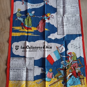 Puede incluir: Una toalla de cocina colorida con un calendario para el año 1789-1989. La toalla presenta una bandera francesa y una escena de personas bailando en círculo. El texto "Les Calissons d'Aix" está impreso en la toalla.