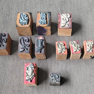 May include: A collection of vintage letterpress stamps. The stamps are made of wood and feature ornate, stylized letter designs in black and gray. Some stamps have a pink or purple background. The stamps are arranged on a gray surface.
