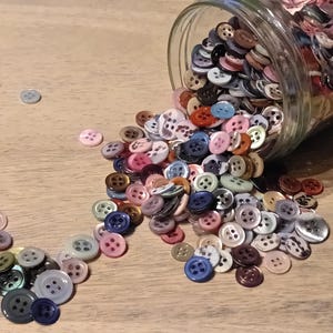 May include: A clear glass jar tipped over, spilling a variety of colorful buttons onto a light-colored wooden surface. The buttons are in shades of pink, blue, orange, and white, with four holes for sewing.