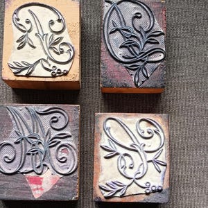 May include: Four vintage letterpress printing blocks. Each block features a different ornate letter design, including the letters A, C, M, and S. The letters are in a dark color, with floral and scrollwork details. The blocks are mounted on wooden bases.