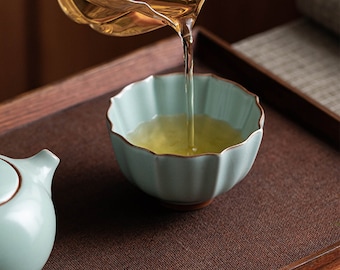 May include: A light green ceramic teacup with a fluted rim is being filled with light yellow tea from a clear glass teapot. The teacup and teapot are on a brown wooden tray.