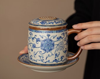 Puede incluir: Una taza de té de cerámica blanca con un diseño floral azul y blanco. La taza tiene una tapa y un asa. La taza está sentada sobre un platillo a juego.