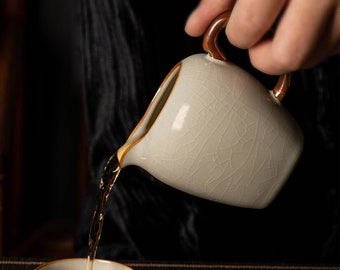 May include: A white ceramic teapot with a brown handle is pouring tea into a white ceramic teacup. The teapot has a crackled glaze finish.