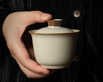 May include: Cream-colored ceramic gaiwan tea cup with a matching lid. The cup and lid have a brown rim and a crackle glaze finish. The gaiwan is held in a person's hand against a dark background.