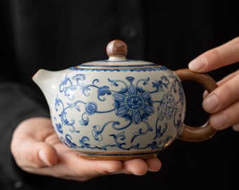 Puede incluir: Una tetera de cerámica blanca con un diseño floral azul y dorado. La tetera tiene un cuerpo redondo y un pico curvo. El asa es marrón.