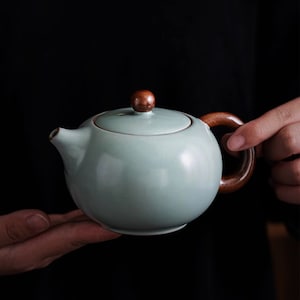 Op de afbeelding: Een lichtgroene theepot van keramiek met een houten handvat en deksel. De theepot heeft een rond lichaam en een klein knopje op het deksel.