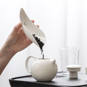 May include: A white ceramic teapot with a rounded body and a small spout is being filled with loose tea from a white scoop. The tea is dark in colour. The teapot sits on a dark tray.