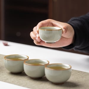 May include: Four small, light green ceramic teacups with brown rims. The cups are arranged in a row on a light brown woven mat. One cup is being held by a hand.