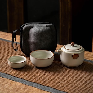 Puede incluir: Un juego de té de cerámica blanca con una tetera, una taza de té pequeña y una taza de té grande. El juego está sobre una estera tejida marrón y viene con una funda de transporte gris.