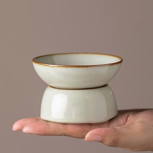 May include: A white ceramic teacup with a brown rim, stacked on top of a similar but slightly larger cup. The cups are both glazed and have a smooth, polished finish.