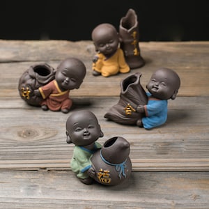 May include: Four small brown ceramic figurines of smiling monks holding brown ceramic pots. The pots have gold Chinese characters on them. The monks are wearing different coloured robes.