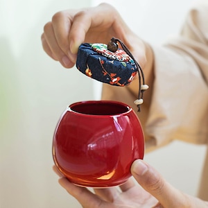 Könnte beinhalten: Eine rote Keramikdose mit runder Form und einem Stoffdeckel mit Blumenmuster. Der Deckel hat einen kleinen Holzknopf und eine braune Schnur.