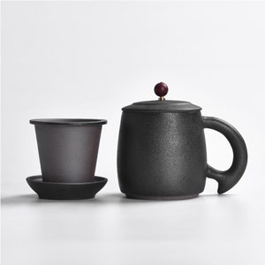 May include: A black ceramic teacup with a lid and a handle. The cup has a textured surface and a small wooden knob on the lid. The cup is sitting on a white surface.