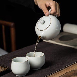 Puede incluir: Una tetera de cerámica blanca con tapa vierte té en dos tazas de té de cerámica blanca. La tetera tiene un borde dorado alrededor de la tapa y el pico. Las tazas de té están sobre una mesa de madera marrón.