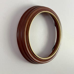 May include: A circular picture frame featuring a smooth, dark brown wooden outer border with a stepped profile and a thin, metallic gold-toned inner rim. The frame is empty, showcasing a hollow center, and is viewed from a slight side angle.