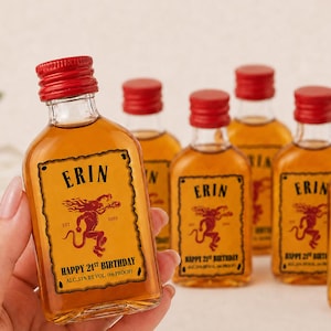 May include: A small glass bottle filled with amber liquid, featuring a red screw cap and a rectangular label. The label displays the name ERIN, a red devil illustration, the text EST 2003, HAPPY 21ST BIRTHDAY, and ALC. 33% BY VOL. (66 PROOF).