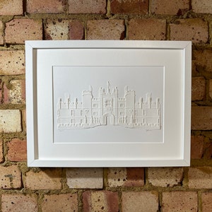 May include: A white framed print of a white paper cut-out of a building. The building is a large, multi-story structure with a central archway and multiple windows. The print is on a brick wall background.